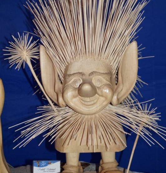 Eine hölzerne Trollfigur mit stacheligem Kopf und lächelndem Gesicht steht auf einer Holzbasis. Die Figur hält einen Stock mit einem Löwenzahn oben. Die Basis hat die Worte Original RS-Troll. Eine kleine Karte mit Text ist auf der Basis platziert.