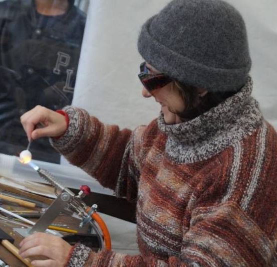 Eine Frau arbeitet in einer Werkstatt an einem Glasprojekt mit einer Fackel. Sie trägt eine graue Mütze, eine Sonnenbrille und einen gestrickten Pullover.