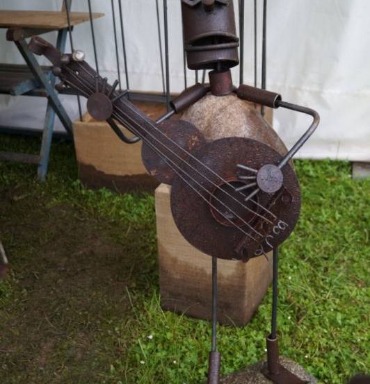 Eine rostige Metallskulptur eines Mannes, der Gitarre spielt, steht auf einem Stein in einem Grasbereich. Hinter ihm befinden sich ein Holzkasten und eine Bank.