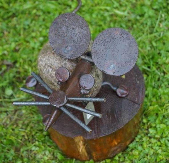 Eine Maus-Skulptur aus Metall und Holz auf einer grasbewachsenen Oberfläche. Sie zeigt einen Mauskopf mit runden Ohren und einem hölzernen Körper.