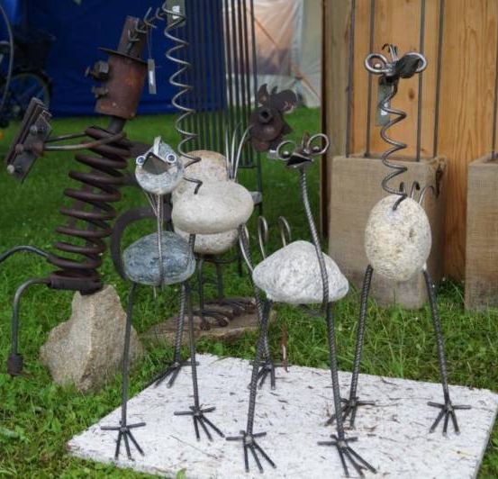 Eine Gruppe von Metallskulpturen von Vögeln steht auf einer weißen Oberfläche im Gras. Jede Skulptur besteht aus Metallstangen und Steinen. Der Hintergrund zeigt eine Holzwand und ein blaues Zelt.