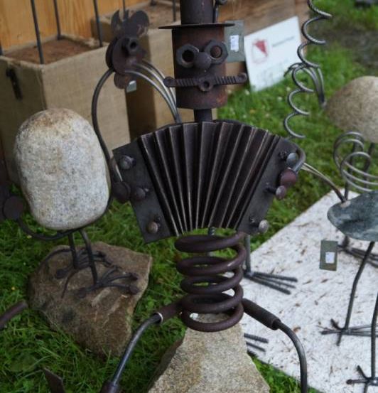 Eine Akkordeon-Skulptur steht auf einem Felsen, mit einer Vogelstatue auf der linken Seite. Dahinter ist eine weiße Karte auf dem Boden platziert. Links steht eine Holzstruktur, und andere Skulpturen sind rechts.