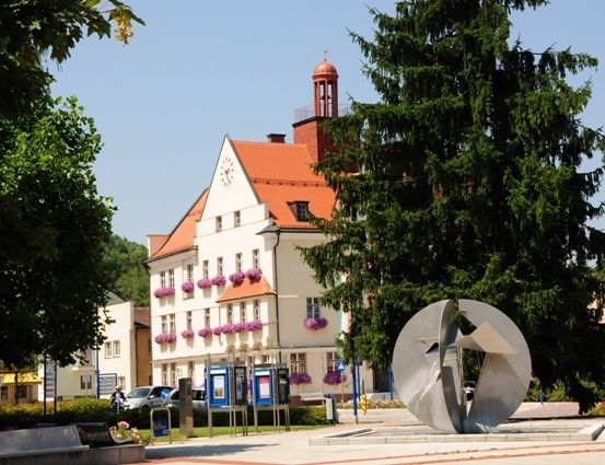 Ein Stadtplatz mit einem großen weißen Gebäude, rotem Dach und Turm. Eine Metallskulptur davor. Bäume und Pflanzen drumherum.