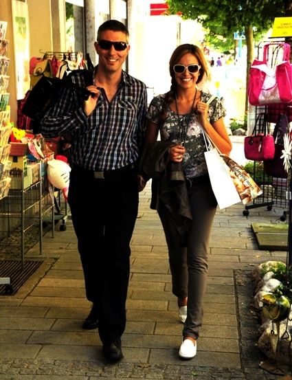 Ein Mann und eine Frau gehen auf einem gepflasterten Bürgersteig. Sie lächeln und halten Händchen. Sie tragen Sonnenbrillen. Der Mann trägt ein kariertes Poloshirt.