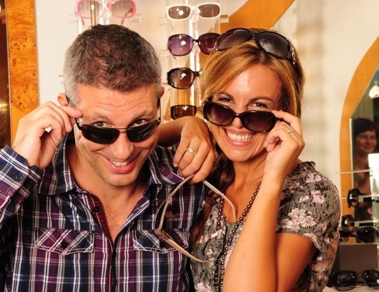 Ein Mann und eine Frau probieren Sonnenbrillen vor einem Spiegel an und lächeln. Der Mann trägt ein kariertes Hemd, und die Frau trägt ein Blumentop. Hinter ihnen hängen Sonnenbrillen an einem Ständer.