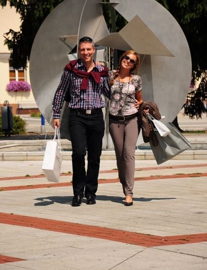 Ein Mann und eine Frau gehen mit Einkaufstaschen auf einem Bürgersteig in der Nähe einer großen abstrakten Skulptur.