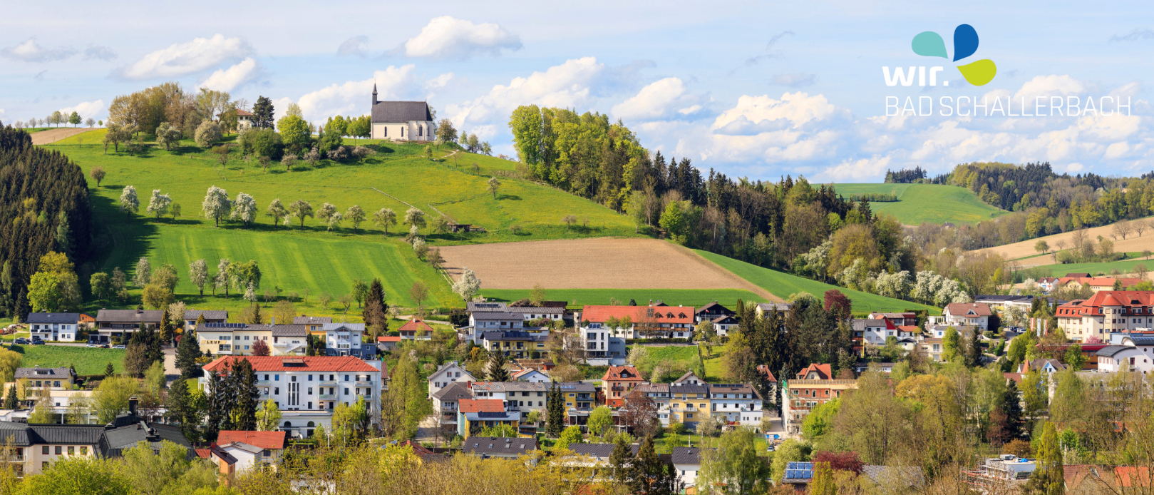 Ein Dorf mit einer Kirche auf einem Hügel, umgeben von üppigen grünen Feldern und Bäumen.