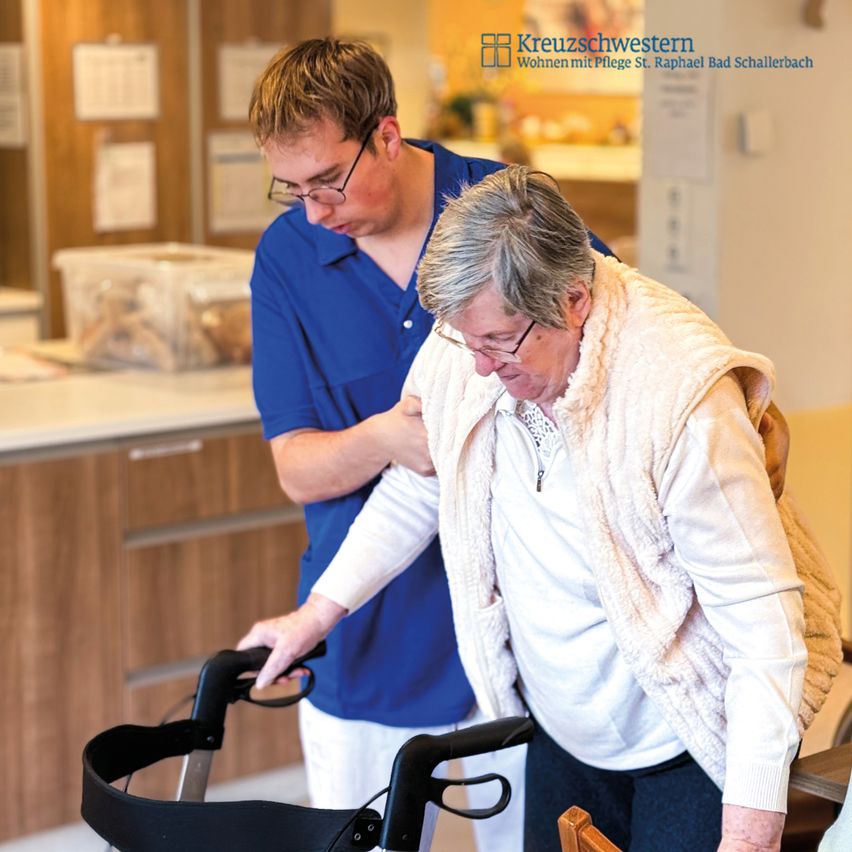 Eine ältere Frau wird von einem Mann unterstützt, während sie einen Gehstock benutzt. Der Hintergrund deutet auf eine Pflegeeinrichtung hin, mit einer modernen Küche im Hintergrund.