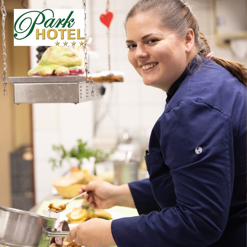 Eine lächelnde Frau in einer blauen Kochuniform kocht am Herd, mit einem Park Hotel Logo über ihr.