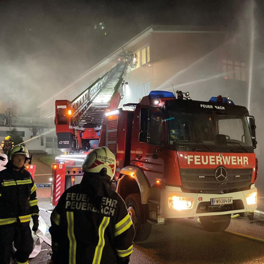 Feuerwehrleute in Schutzkleidung stehen vor einem Feuerwehrwagen. Die Leiter des Wagens ist ausgefahren und Wasser spritzt aus ihr. Das Gebäude hinter ihnen ist in Rauch gehüllt.