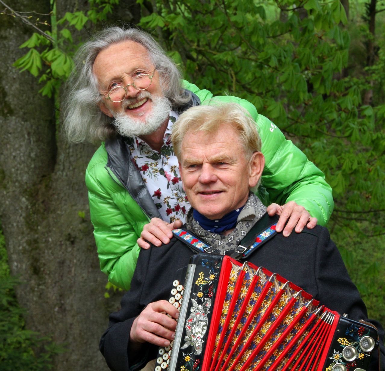 Zwei Männer stehen in einem Wald. Einer spielt Akkordeon und der andere steht hinter ihm mit der Hand auf seiner Schulter.