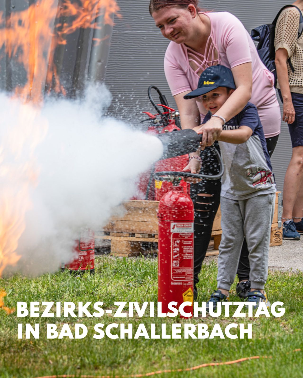 Ein junger Junge benutzt einen Feuerlöscher mit der Hilfe eines Erwachsenen auf einem Rasen mit einem Feuer im Hintergrund. Die Veranstaltung findet in Bad Schallerbach statt.