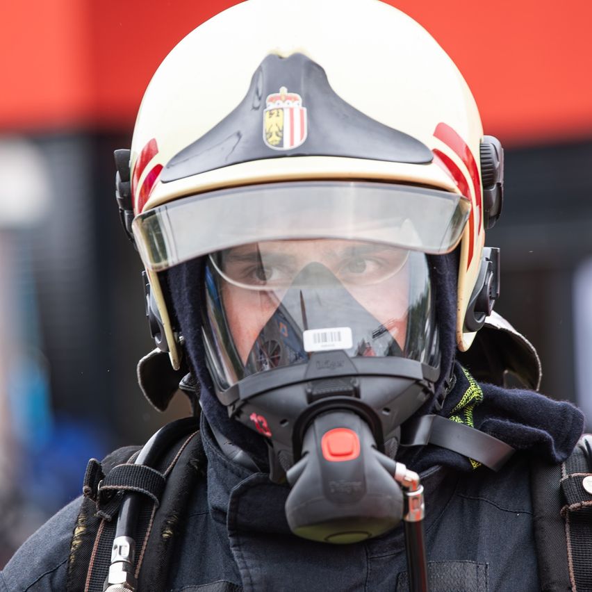 Ein Feuerwehrmann in voller Ausrüstung, einschließlich eines Helms mit sichtbarem Logo, steht bereit. Er trägt eine Maske und ein Atemgerät.