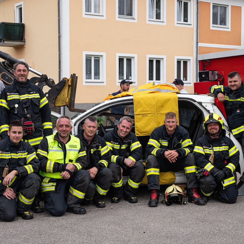 Eine Gruppe Feuerwehrleute posiert für ein Foto vor einem Auto und einem Feuerwehrauto, mit einem Gebäude im Hintergrund.