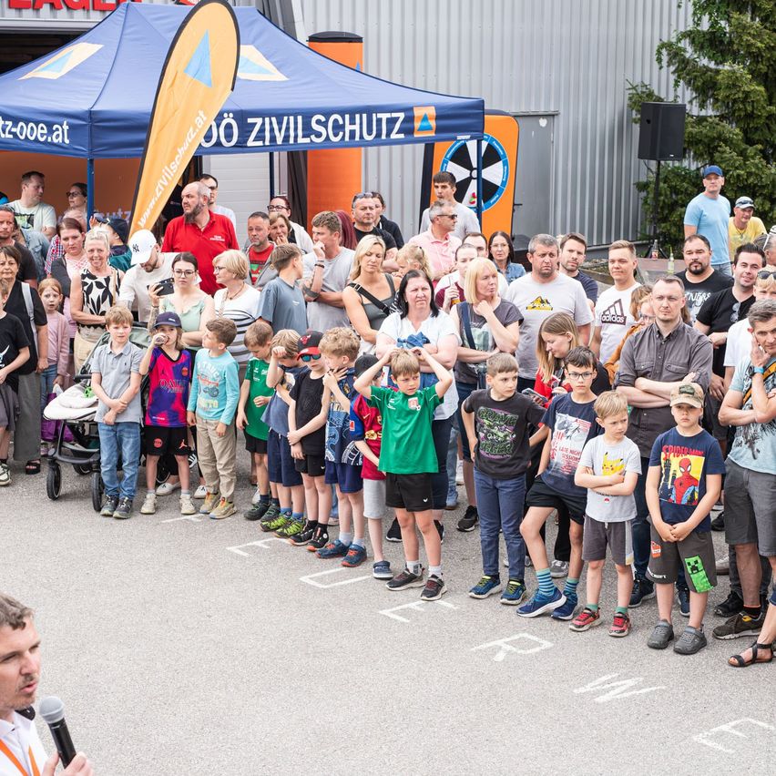 Eine Menschenmenge, darunter auch Kinder, steht in einer Reihe und beobachtet einen Mann, der in ein Mikrofon spricht. Ein Zelt mit einem Banner steht hinter ihnen.