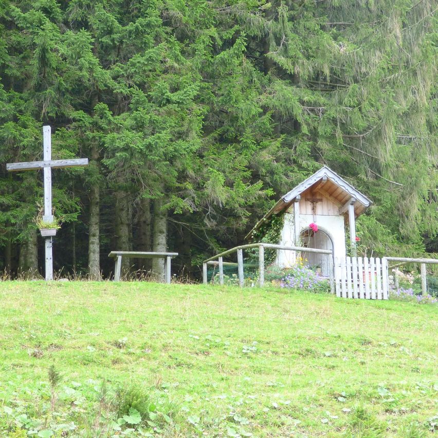 Eine kleine Holzkapelle mit einem Kreuz und einer Bank steht in einem von hohen Bäumen umgebenen Grasbereich.