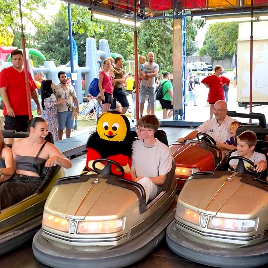 Mehrere Menschen fahren Achterbahnen in einem Vergnügungspark. Ein Auto hat eine Maskottchen darauf. Andere schauen von den Seitenlinien zu.