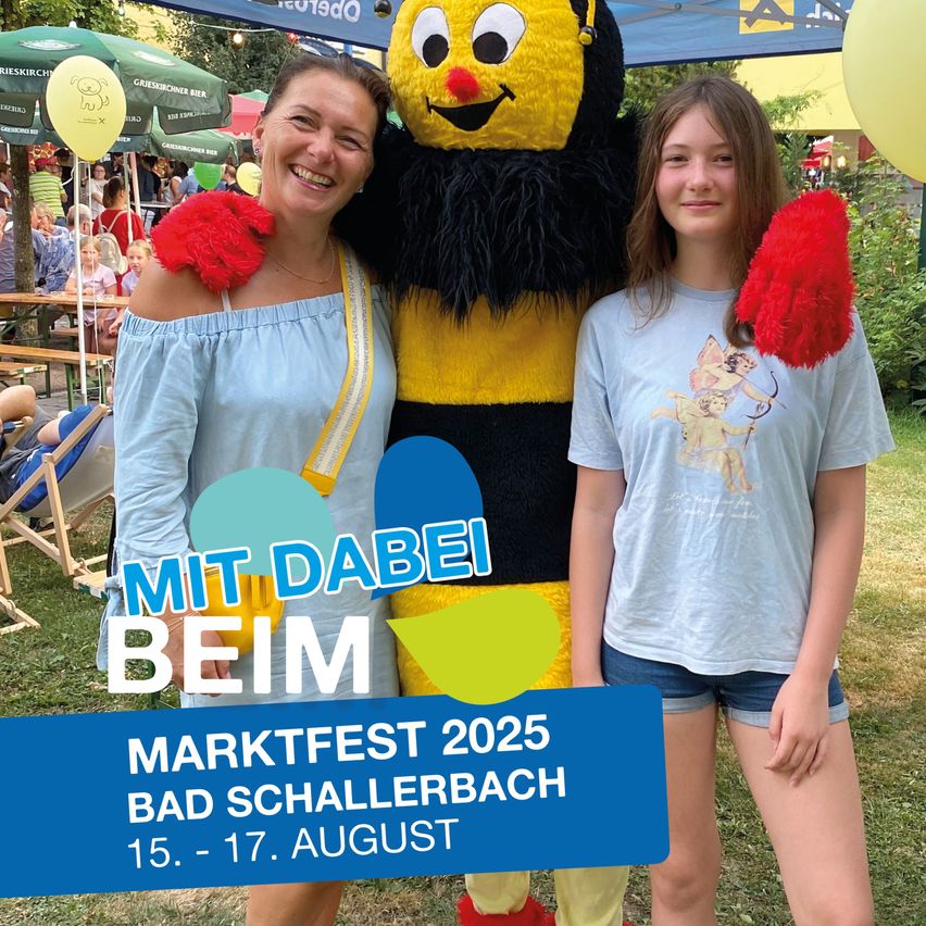 Zwei Frauen stehen neben einer Maskottchen, einer großen gelben und schwarzen Biene, auf einem Festival. Eine Frau trägt ein blaues Shirt, die andere ein graues Shirt mit einem Bogen-und-Pfeil-Design. Hinter ihnen ist ein Markt mit Menschen, Zelten und Luftballons.