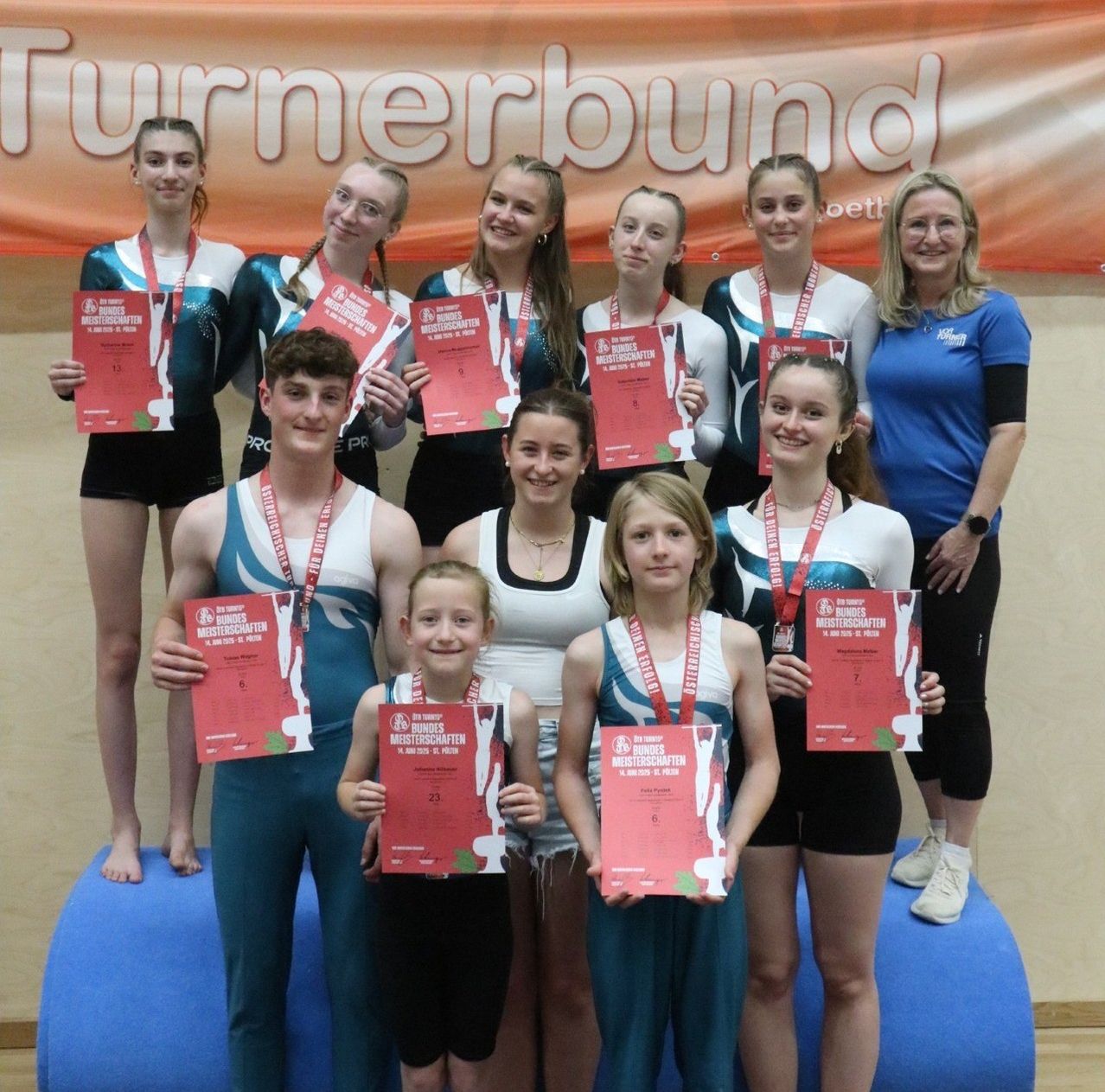 Eine Gruppe junger Athleten und Trainer posiert mit Medaillen und Urkunden auf einer blauen Matte vor einem Hintergrund mit der Aufschrift 'Turnerbund'.