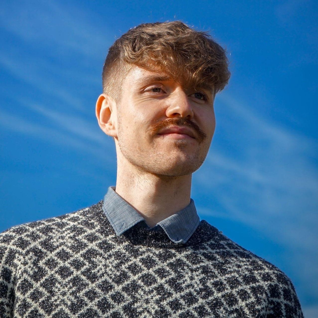 Ein Mann mit einem Schnurrbart, der einen karierten Pullover und ein Hemd mit Kragen trägt, steht vor einem blauen Himmel.
