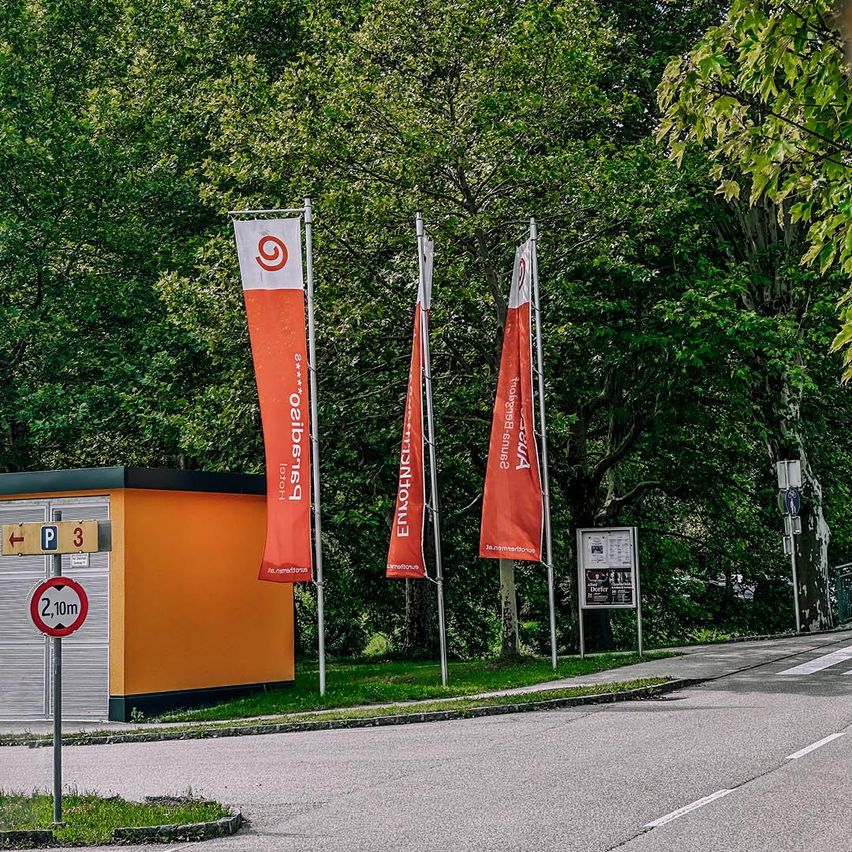 Ein Parkplatzschild und drei Banner sind vor einem Gebäude mit Bäumen und einer Straße im Hintergrund zu sehen.