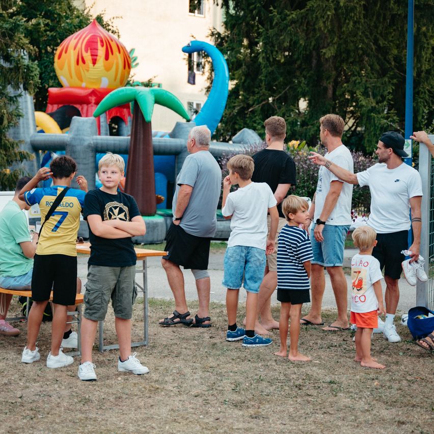 Eine Gruppe von Menschen steht in einem Außenbereich mit aufblasbaren Spielen im Hintergrund. Einige sind Erwachsene und andere sind Kinder, alle in Freizeitkleidung.
