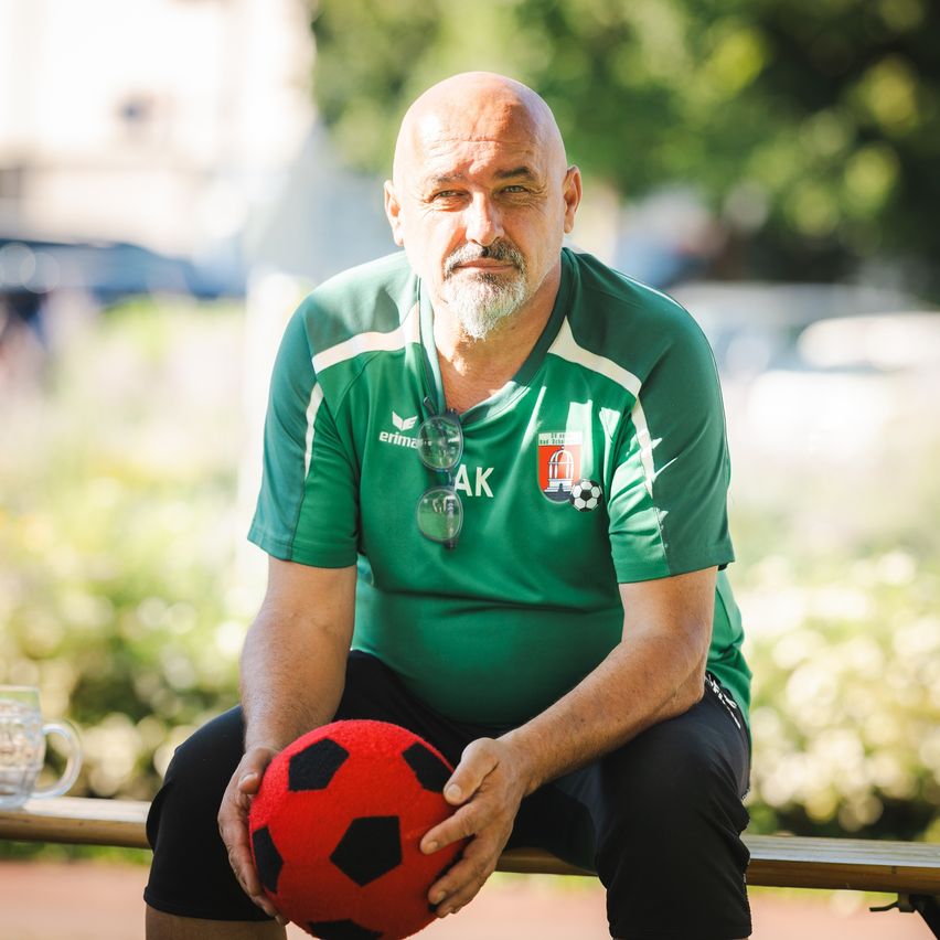 Ein kahlköpfiger Mann mit Bart sitzt auf einer Bank und trägt ein grünes Trikot mit weißen Streifen und den Buchstaben AK. Er hält einen roten Fußball und hat eine Sonnenbrille um den Hals.