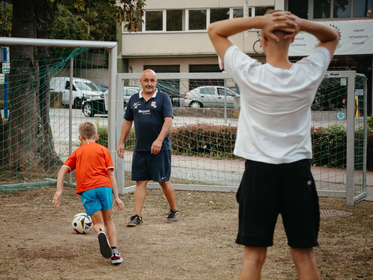 Ein Mann in einem blauen Trikot und Shorts lehrt einen jungen Jungen in Orange, einen Fußball zu schießen, in der Nähe eines Tores. Ein weiterer Junge in Weiß beobachtet von hinten.