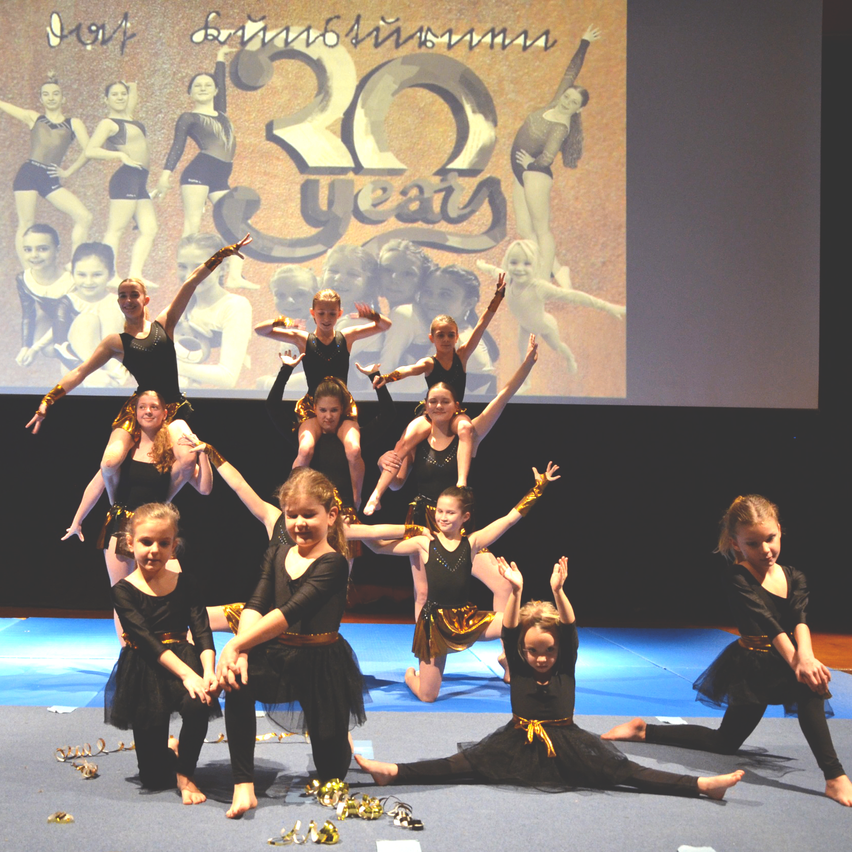 Eine Gruppe junger Mädchen in schwarzen und goldenen Kostümen führt Akrobatik auf einer Bühne vor, auf der ein großer Bildschirm mit Bildern von Tänzern im Hintergrund steht.
