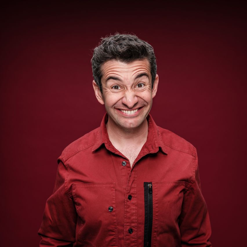 Ein Mann mit kurzen, dunklen Haaren trägt ein rotes Hemd und lächelt in die Kamera. Er hat Falten um die Augen und steht vor einem roten Hintergrund.