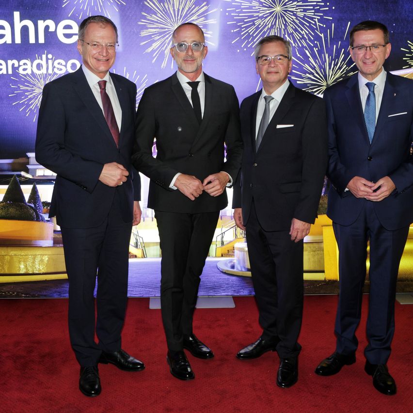 Vier Männer in Anzügen stehen lächelnd auf einem roten Teppich, mit Feuerwerk im Hintergrund.
