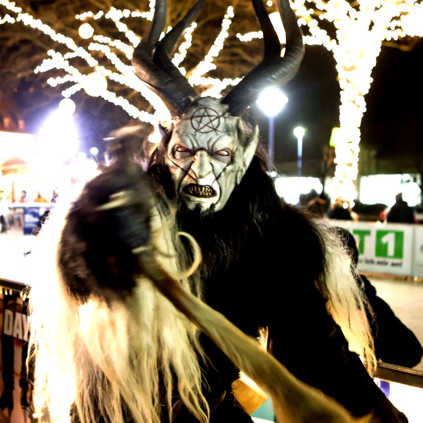 Eine Person in einer gruseligen Maske mit Hörnern steht nachts draußen. Die Maske hat ein Pentagrammsymbol darauf. Im Hintergrund sind Menschen und Lichter zu sehen.