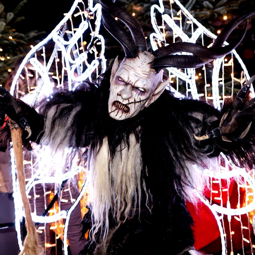 Eine Person in einem hörnertragenden Dämonenkostüm steht unter dekorativen Lichtern, möglicherweise auf einem Weihnachtsmarkt.