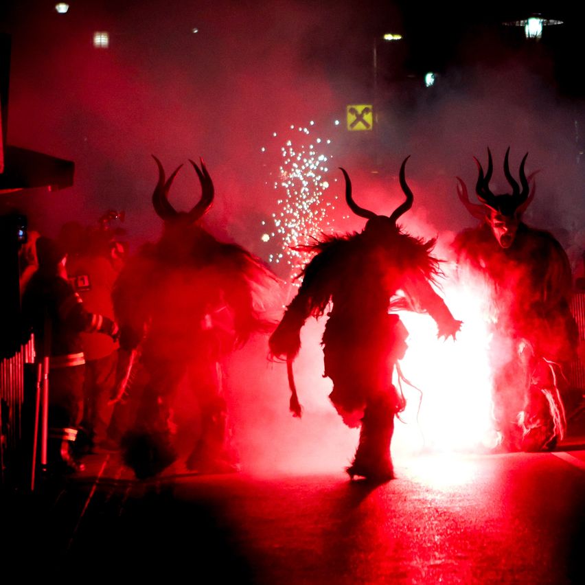 Eine Gruppe von Menschen in Hörnerkostümen tritt in einer rot beleuchteten Umgebung mit Funken in der Luft auf. Ein helles Licht beleuchtet die zentrale Figur.