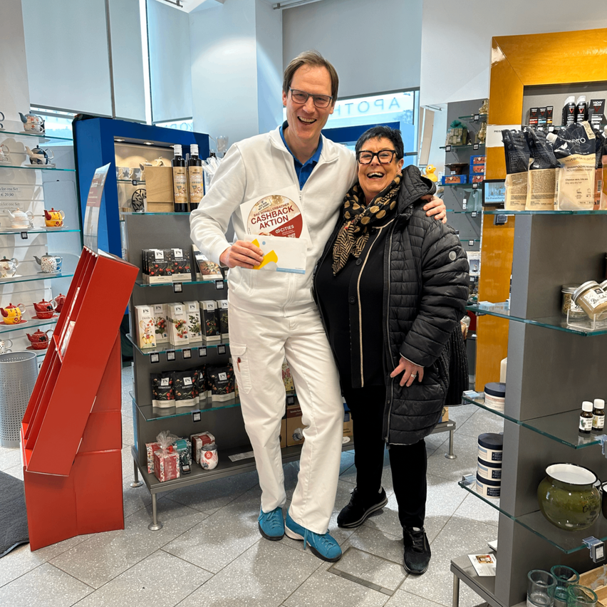 Ein Mann und eine Frau stehen lächelnd in einem Geschäft, der Mann hält ein Schild mit der Aufschrift 'Cashback Aktion'. Sie sind von Regalen mit verschiedenen Produkten umgeben, und der Mann trägt einen weißen Laborkittel und blaue Schuhe, während die Frau eine schwarze Jacke trägt.
