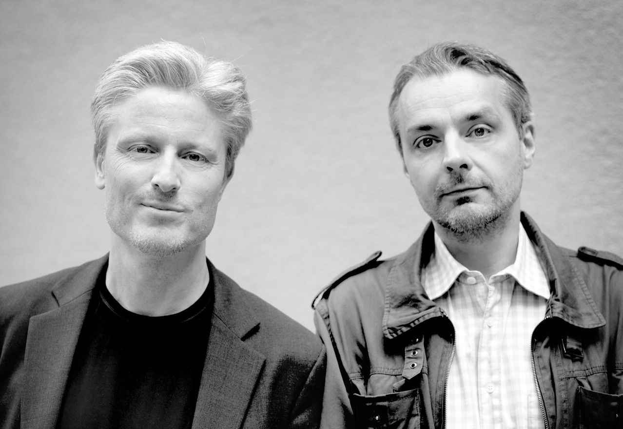 Ein schwarz-weißes Porträt von zwei Männern, die eng beieinander stehen. Ein Mann hat einen Bart und trägt eine Jacke, während der andere Mann kurze Haare hat und ein Hemd trägt.