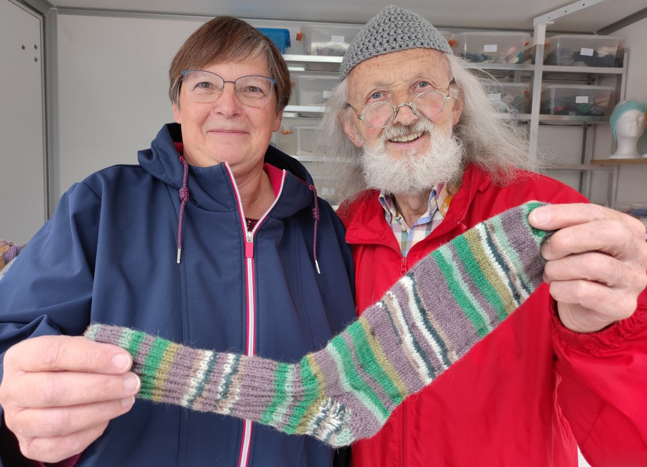 Ein älterer Mann und eine Frau lächeln und halten einen bunt gestreiften Schal in einer Werkstatt.