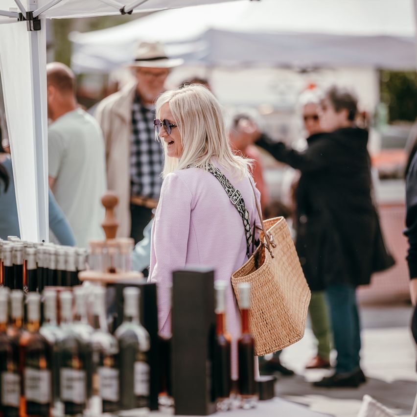 Eine Frau mit blonden Haaren, die eine Sonnenbrille und einen rosa Pullover trägt, lächelt auf einem überfüllten Markt mit Flaschen Schnaps.