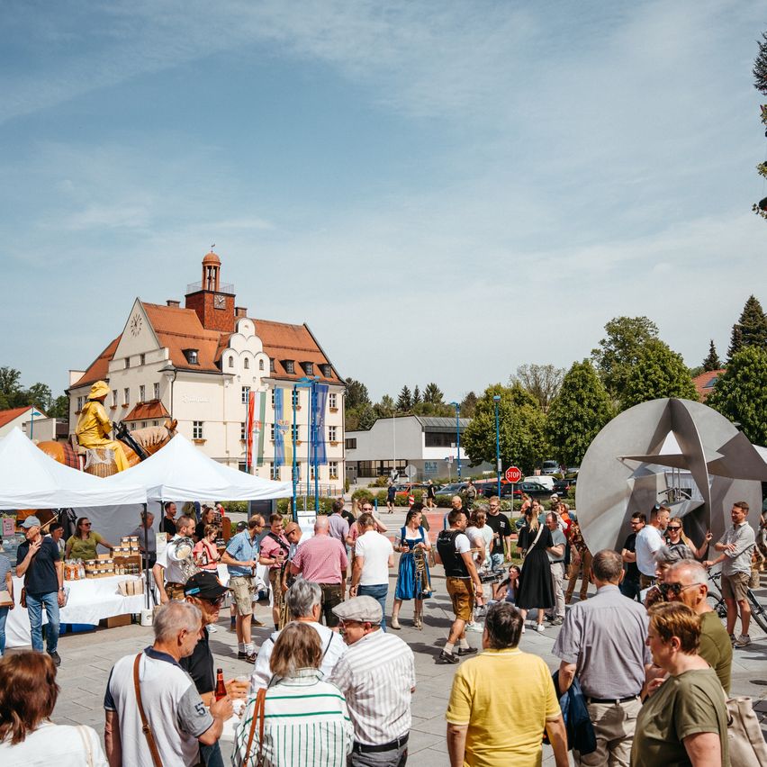 Eine Menschenmenge versammelt sich auf einem Außenplatz, einige sitzen um Tische unter weißen Zelten. Eine große Metallskulptur befindet sich im Hintergrund in der Nähe eines Gebäudes mit Turm und Uhr. Straßenlaternen und Bäume sind ebenfalls zu sehen.