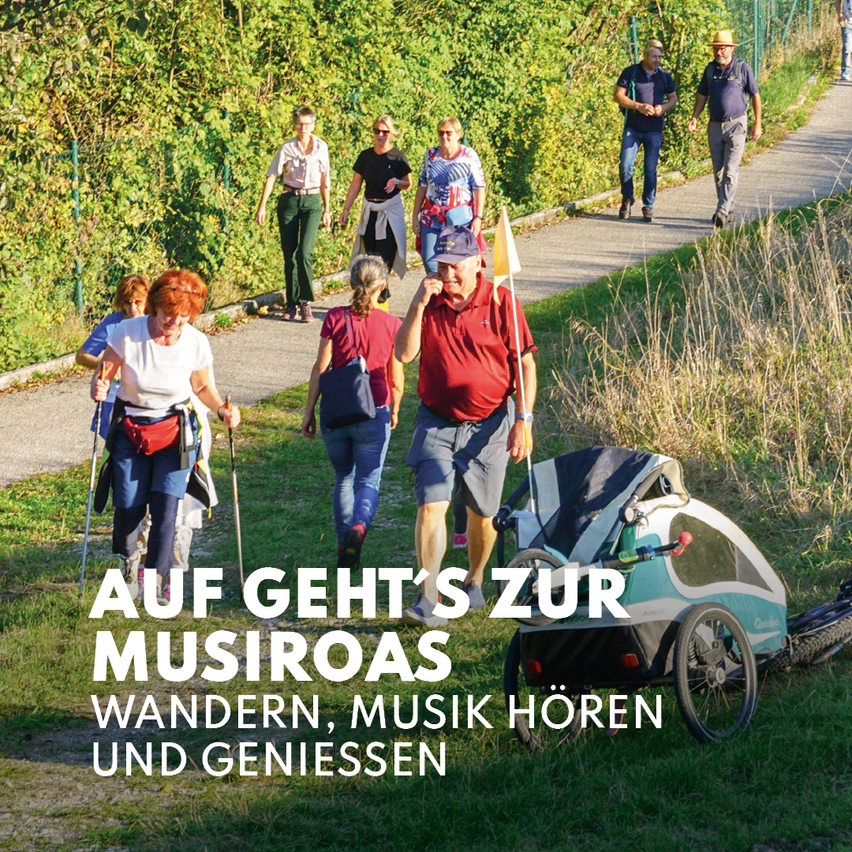 Eine Gruppe von Menschen geht auf einem Pfad mit einem Kinderwagen, ein Mann hält eine Fahne. Der Text lautet 'AUF GEHT'S ZUR MUSIROAS'. Sie sind von Gras und Büschen umgeben.