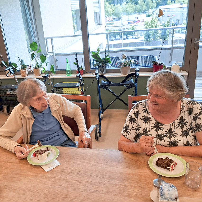 Zwei ältere Frauen sitzen an einem Tisch und essen Kuchen mit Gabeln. Hinter ihnen ein Fenster mit Blick auf einen Parkplatz und Grünflächen.