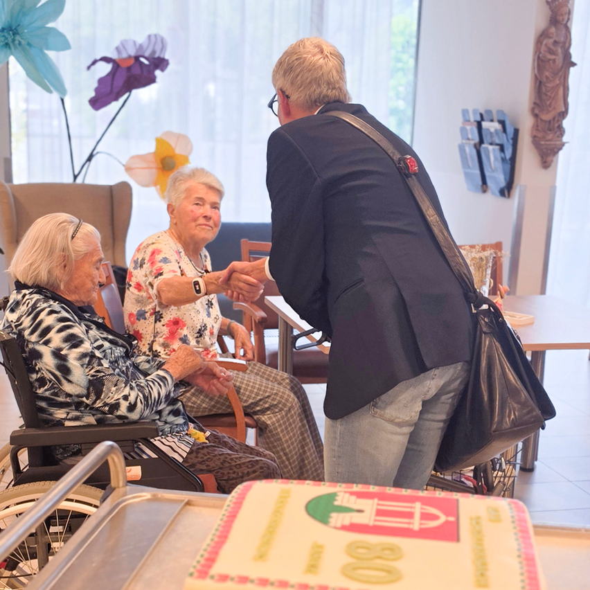 Ein Mann in einem Anzug schüttelt die Hände von zwei älteren Frauen, die in Rollstühlen sitzen, mit einem Kuchen und Blumen im Hintergrund.