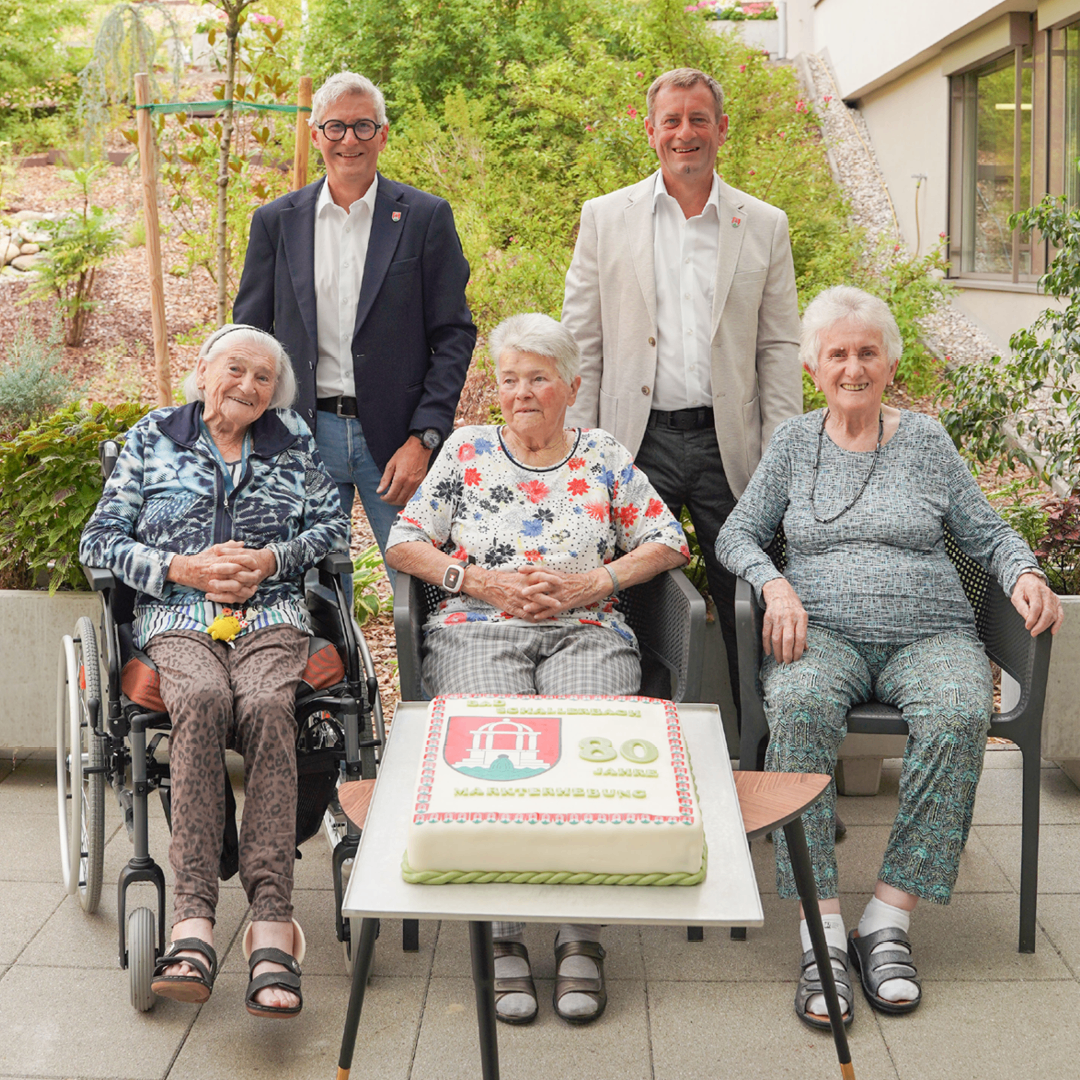Eine Gruppe älterer Frauen und zwei Männer feiern einen Geburtstag mit einem Kuchen vor ihnen. Sie sitzen auf Stühlen und lächeln.