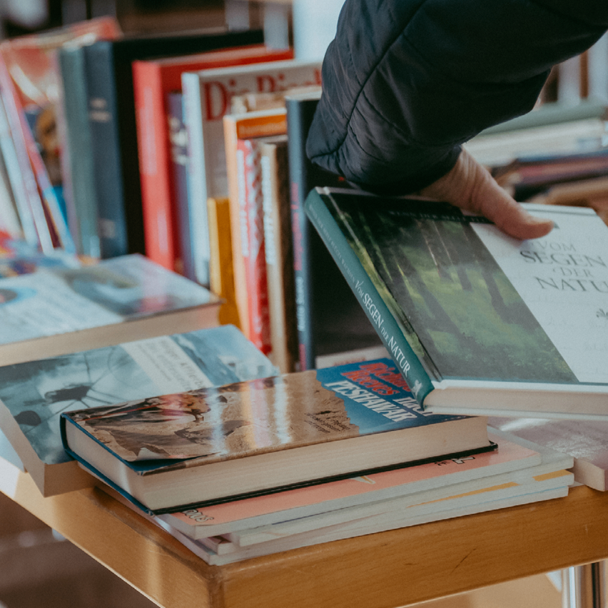 A person is holding a book titled 'Vom Segen der Natur' in front of a table full of books.