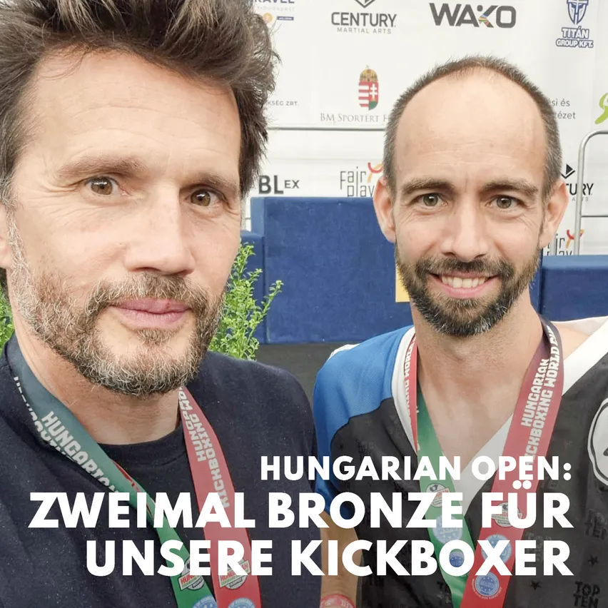 Zwei Männer mit Medaillen um den Hals stehen nebeneinander. Der Mann auf der linken Seite hat einen Bart und trägt eine Medaille um den Hals. Der Mann auf der rechten Seite ist kahl und trägt ebenfalls eine Medaille. Sie lächeln beide und scheinen für ein Foto zu posieren.