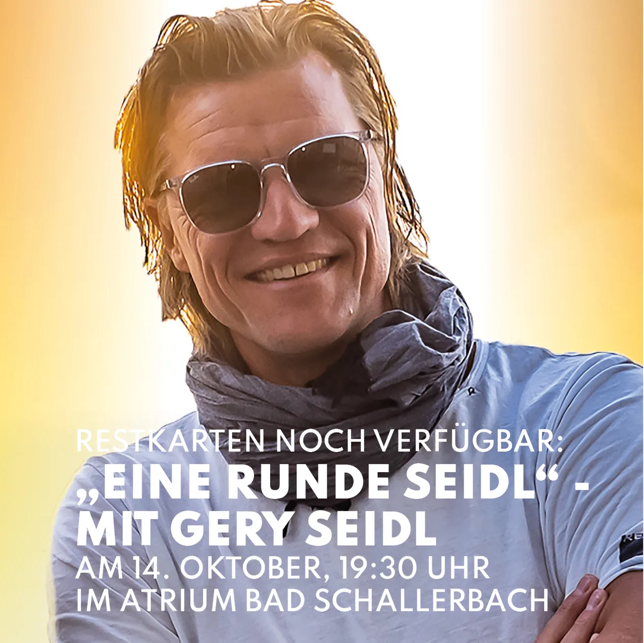 Ein lächelnder Mann mit nassen Haaren und Sonnenbrille trägt ein blaues Hemd und einen Schal.