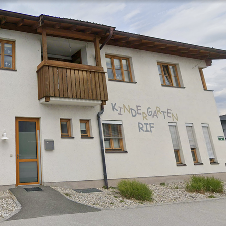 Ein weißes Haus mit hölzernen Balkonen, braunem Zierrat und mehreren Fenstern. Eine hölzerne Tür mit Glasscheiben führt zum Eingang.