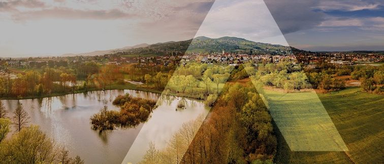 Luftaufnahme einer friedlichen Landschaft mit einem See, üppigen Bäumen und einer Stadt mit Gebäuden, eingebettet zwischen Bergen und Hügeln.