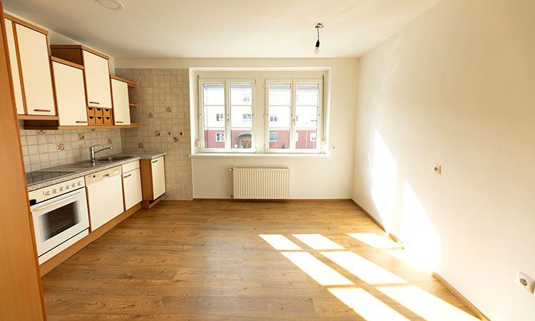 Eine leere Küche mit Holzboden, weißen Schränken, einer Spüle, einem Herd und einem Fenster mit Blick auf ein Gebäude.