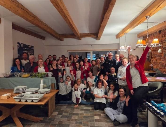 Eine Gruppe von Menschen in einem Raum, einige stehend und einige sitzend, alle lächeln und winken. Es gibt Tische mit Tellern und Essen, und eine Frau in einer roten Jacke hebt ihre Hand.